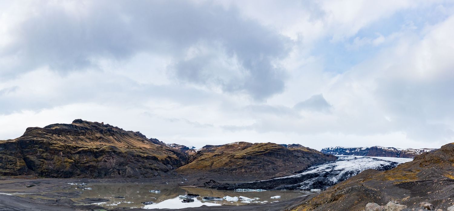 Panoramic view of an Icelandic glacier landscape with black volcanic rock, snow-capped mountains, and cloudy sky.