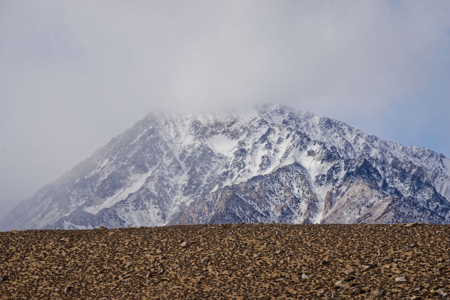 Snow-dusted mountain peak rising above rocky desert terrain under a cloudy sky.