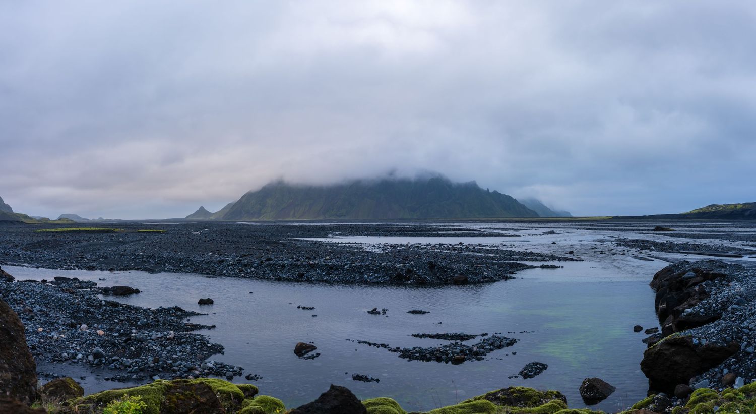Dramatic Icelandic coastal panorama with dark rocky shores, calm reflective waters, and a misty mountain shrouded in clouds.