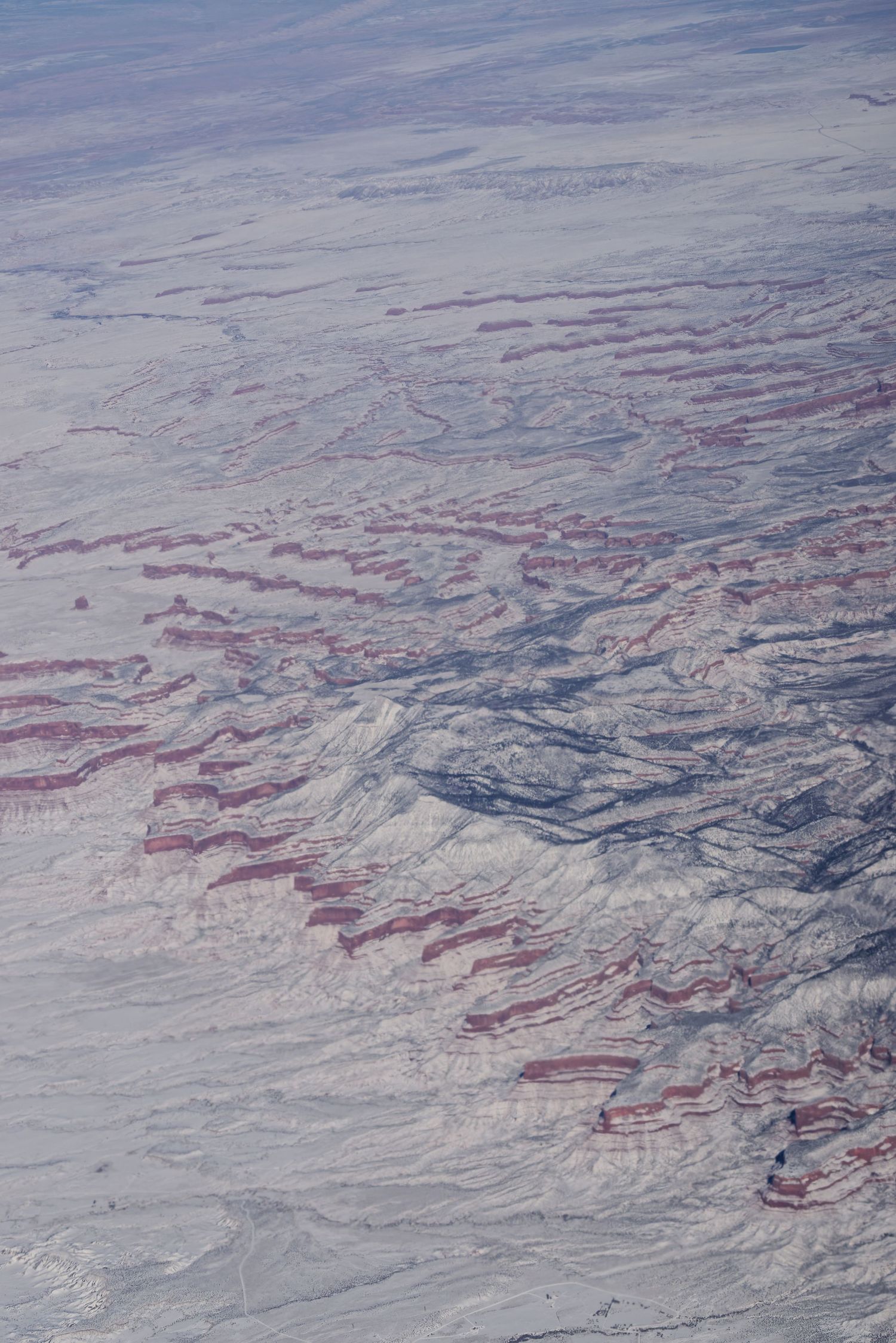Aerial view of snow-covered terrain with red-hued rock formations creating striking patterns across a vast landscape.