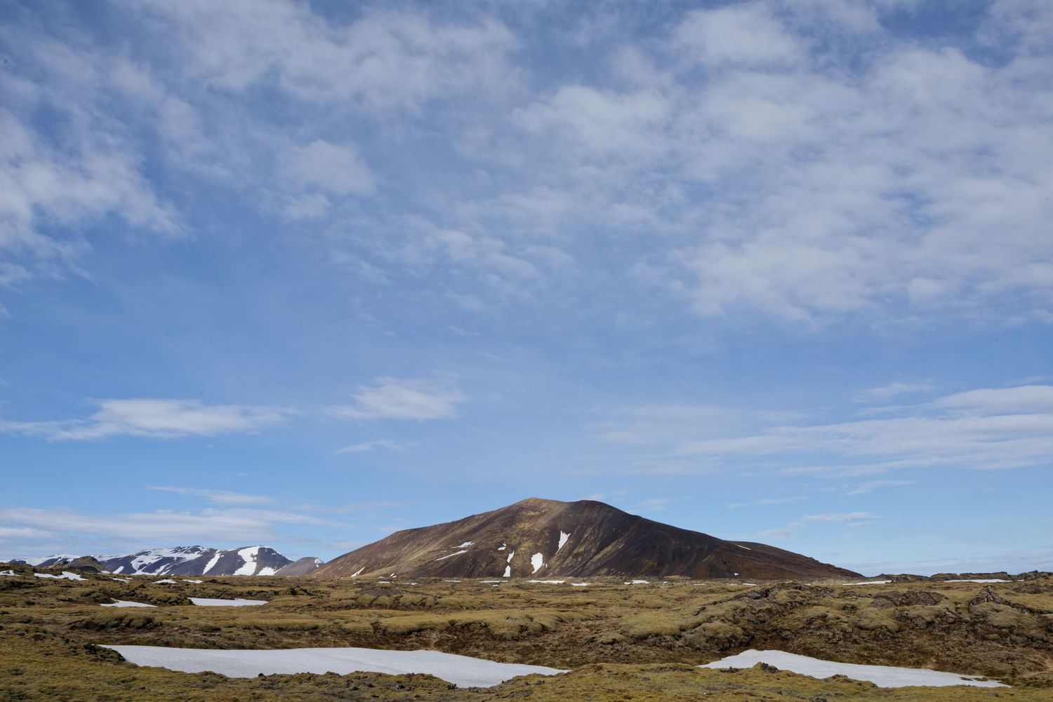 A lone volcanic mountain peak rises above mossy tundra with patches of snow under blue skies.