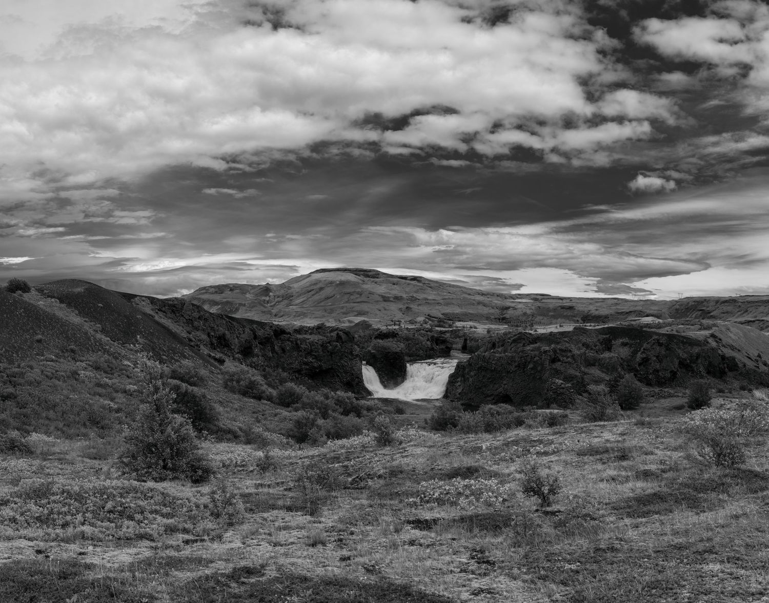 Black and white landscape of Icelandic highlands with waterfall, rolling hills, and dramatic cloudy sky.