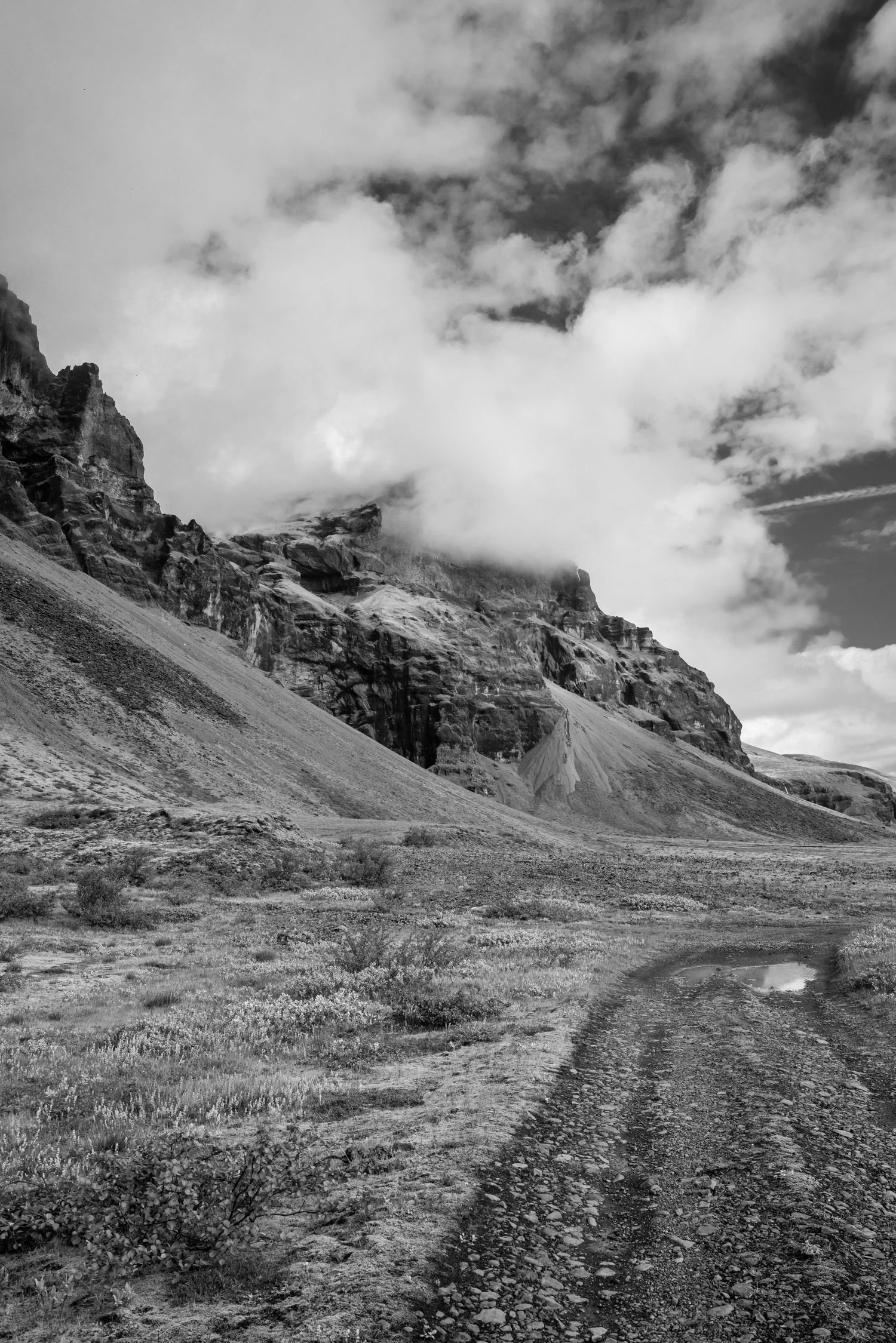 Black and white photo of a rugged dirt road leading toward dramatic rocky mountains with clouds in Iceland.