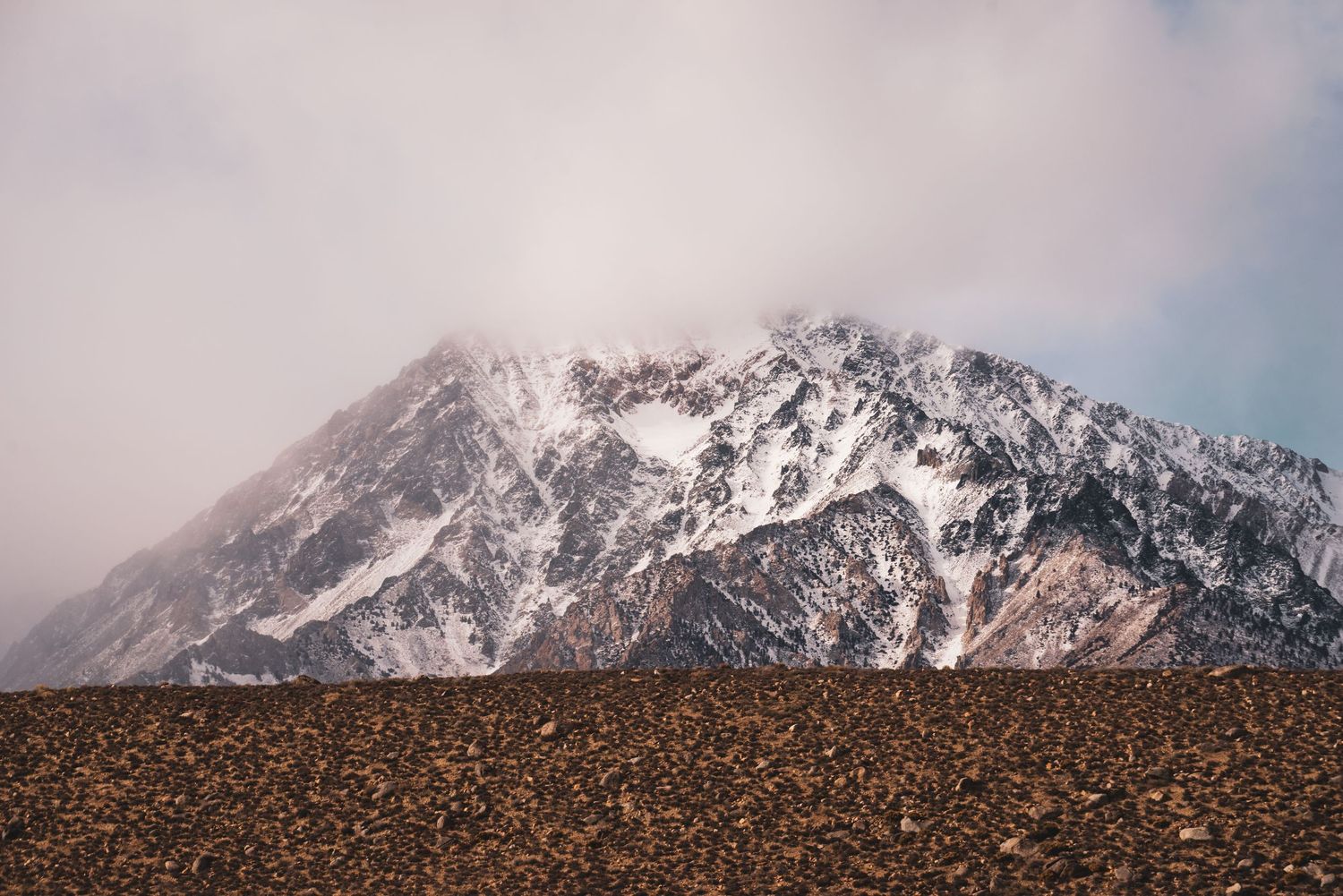 Snow-dusted mountain peak emerging through misty clouds above a rocky desert terrain near Lone Pine California.