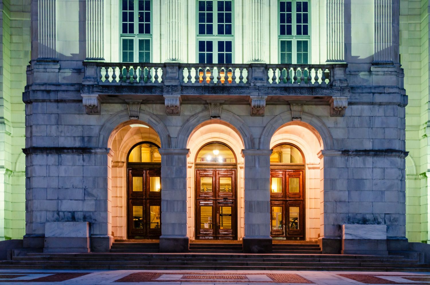 Illuminated neoclassical building facade with three arched doorways, ornate balustrade, and warm golden lighting at night.