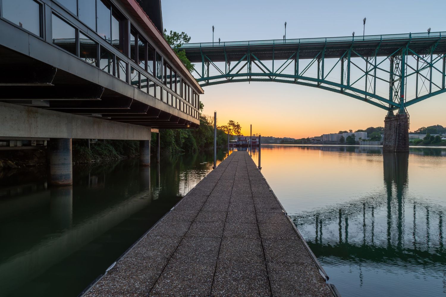 Sunset view of a riverside walkway beneath two bridges reflecting golden light on calm water.