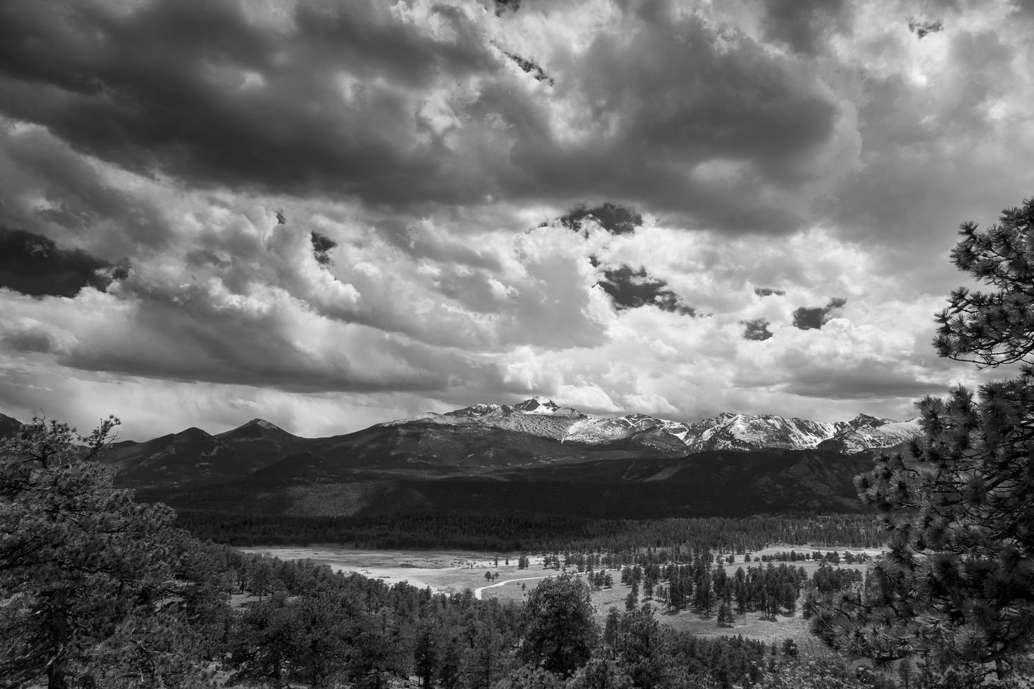 Black and white landscape photo of mountain range with dramatic storm clouds over forested valley and lake.