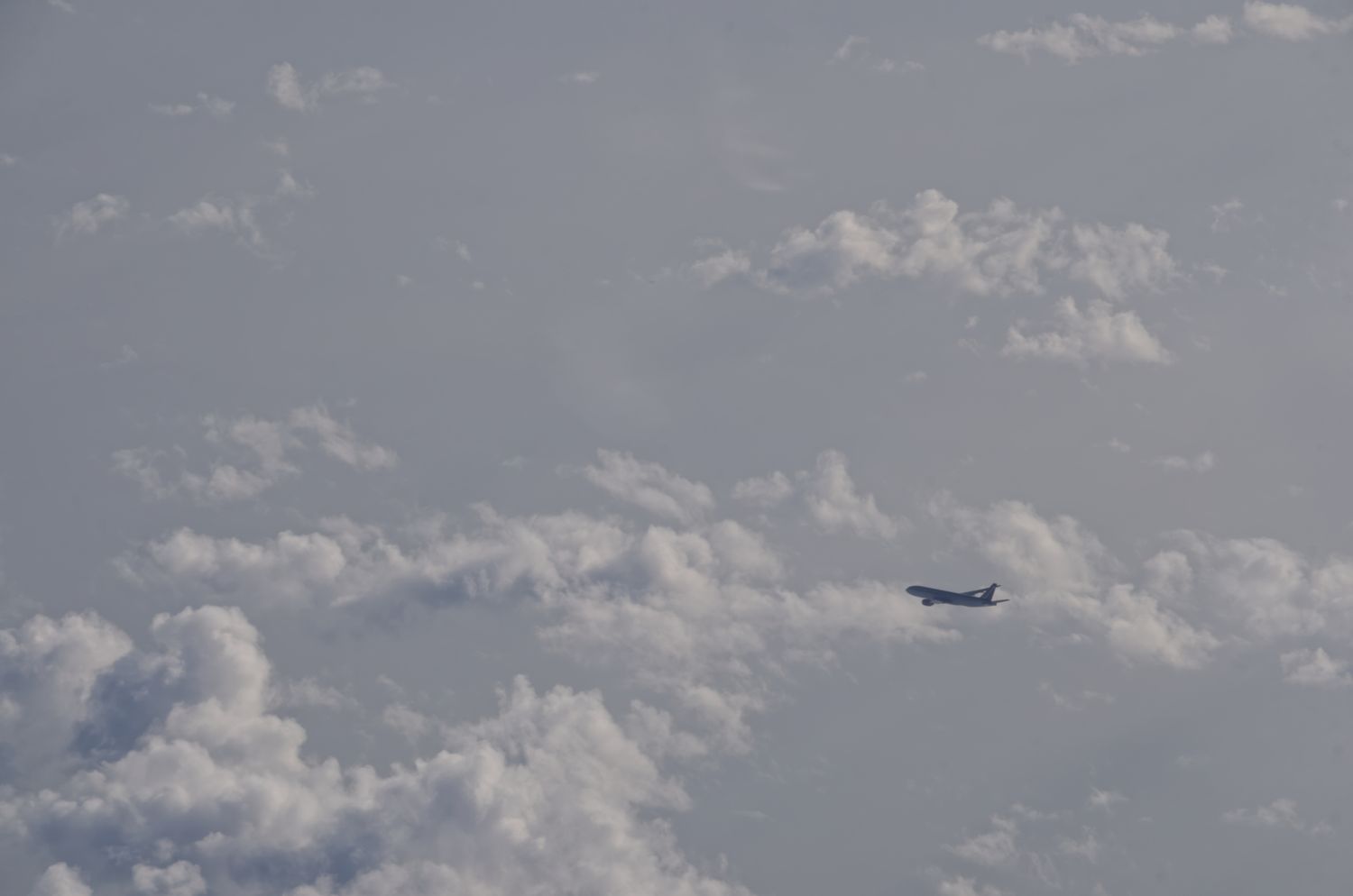 A commercial airplane flies among scattered white clouds viewed from above against a pale blue sky.