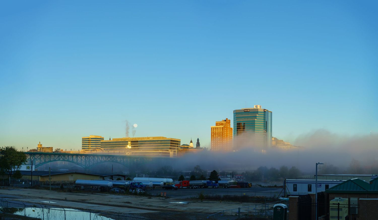 Misty morning cityscape with modern buildings emerging from low fog under a clear blue sky.