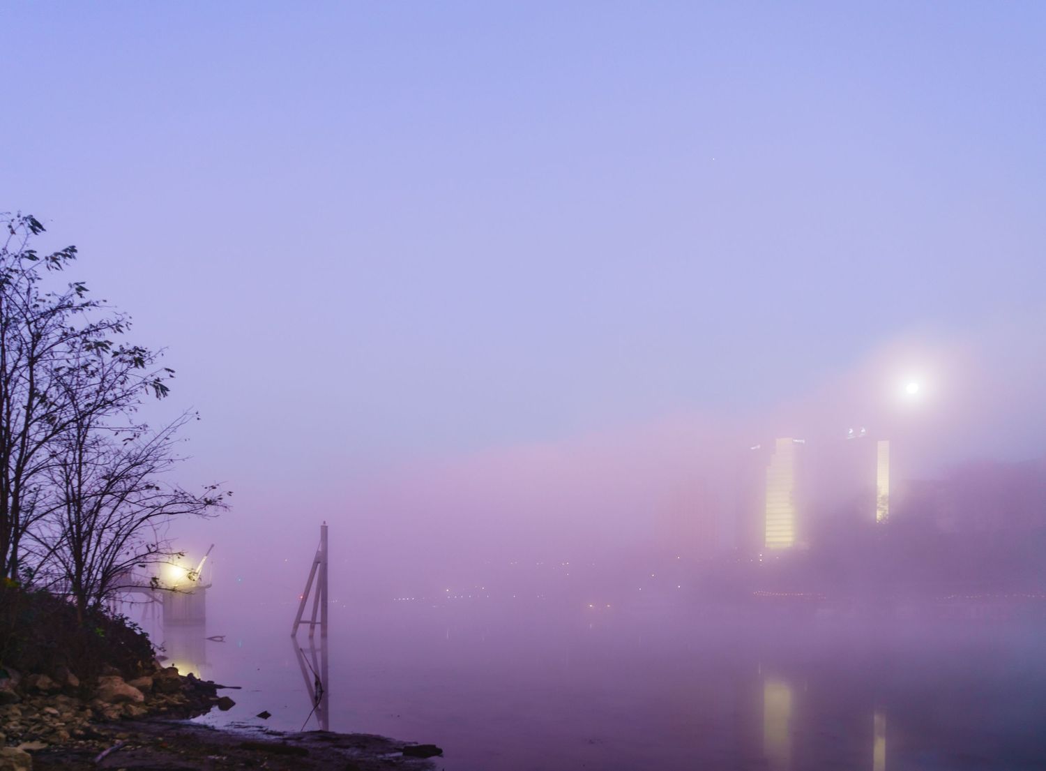 Misty foggy evening over calm river with glowing bridge lights and bare trees in purple twilight atmosphere.