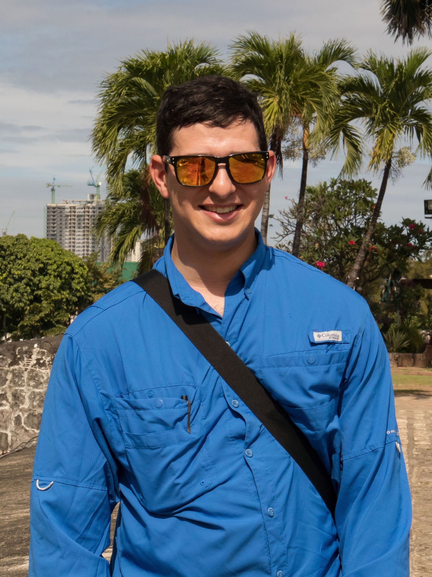 Man in blue Columbia shirt and sunglasses smiling outdoors near palm trees and historic stone wall.