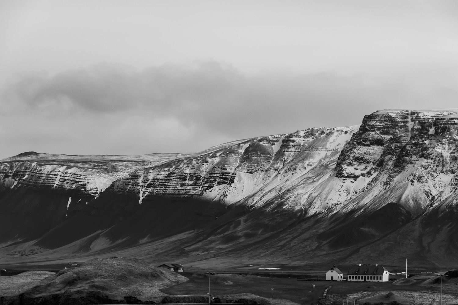 Black and white landscape photo of snow-capped mountains with a small white building in the foreground under overcast skies.