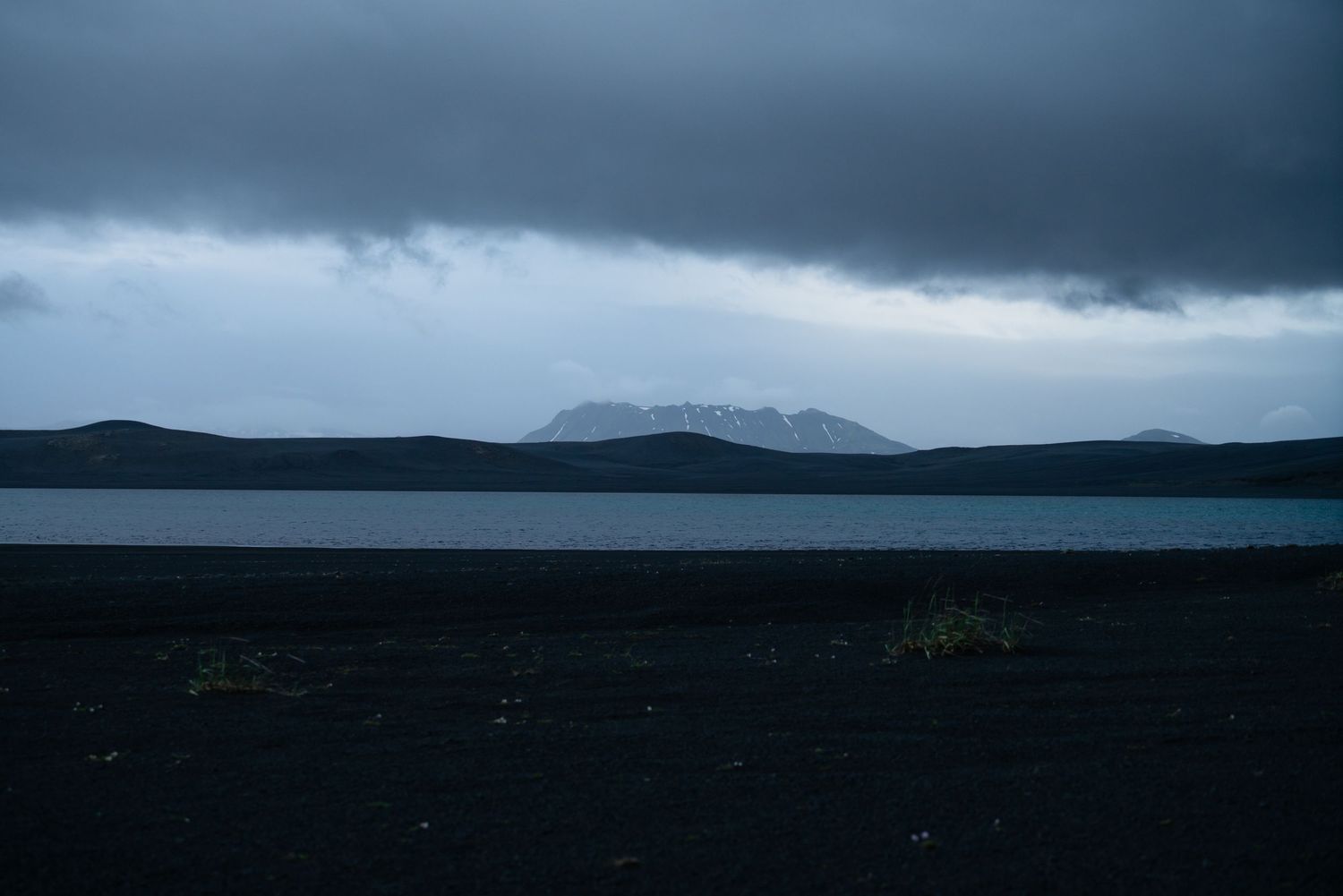 Dark moody landscape with black volcanic shore, calm lake, and snow-capped mountain under stormy clouds in Iceland.