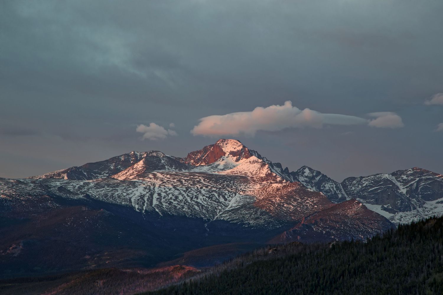 Dramatic sunset over snow-covered mountain peaks with dark storm clouds in the sky.