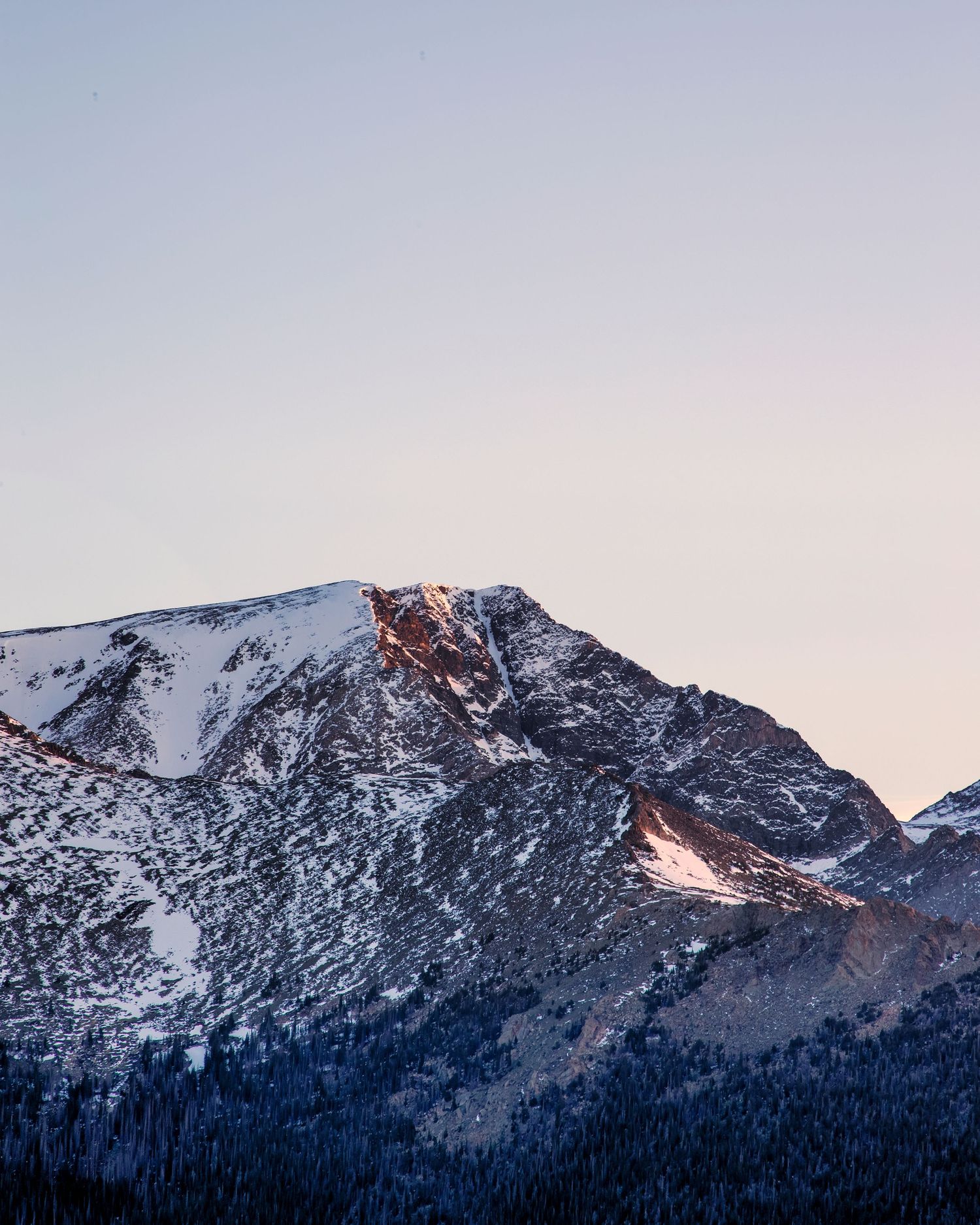 Snow-capped mountain peak with rocky terrain and pine trees at sunset in winter.