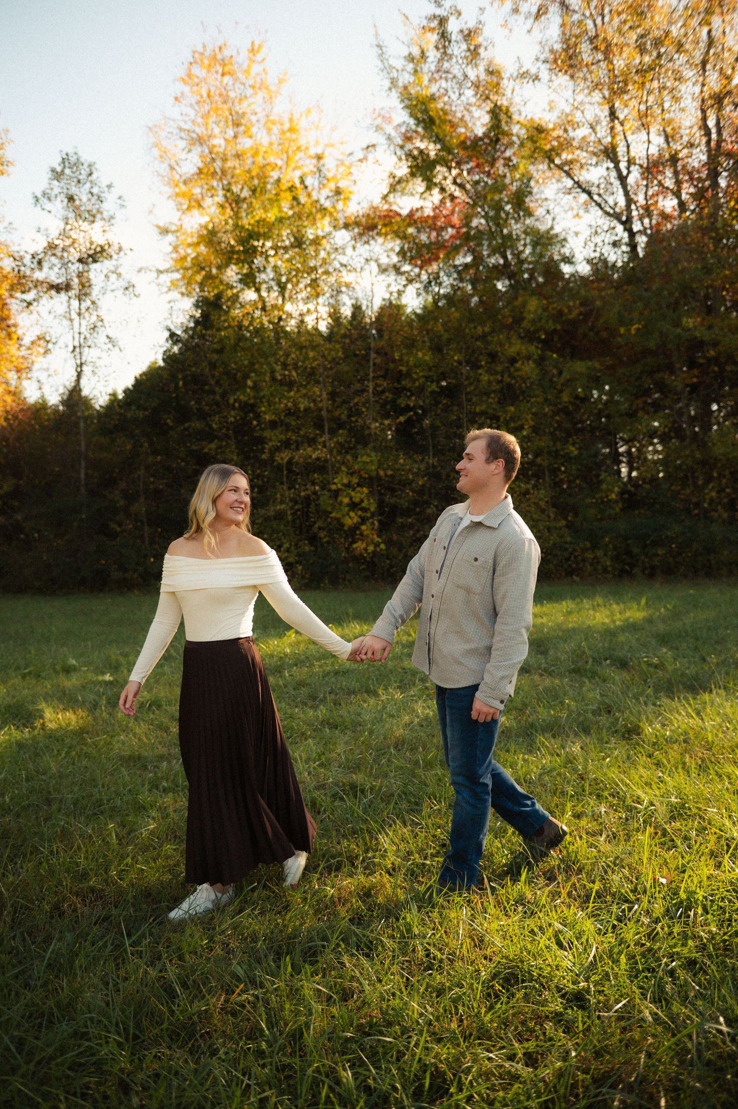 A couple holds hands and walks through a sunlit green field with autumn trees in the background.