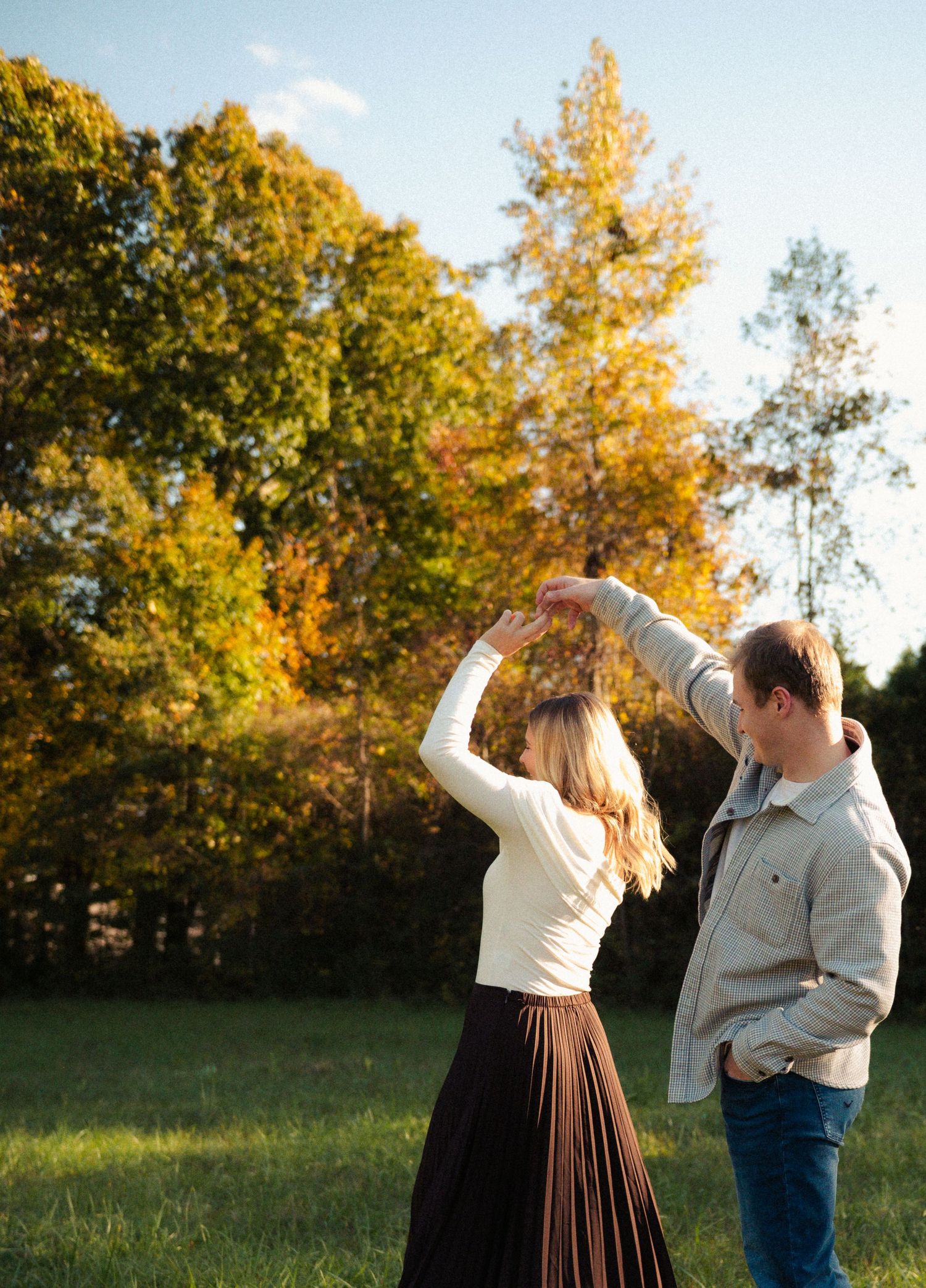 Couple dancing outdoors in autumn park, woman in brown skirt twirling under man's hand amid colorful fall foliage.