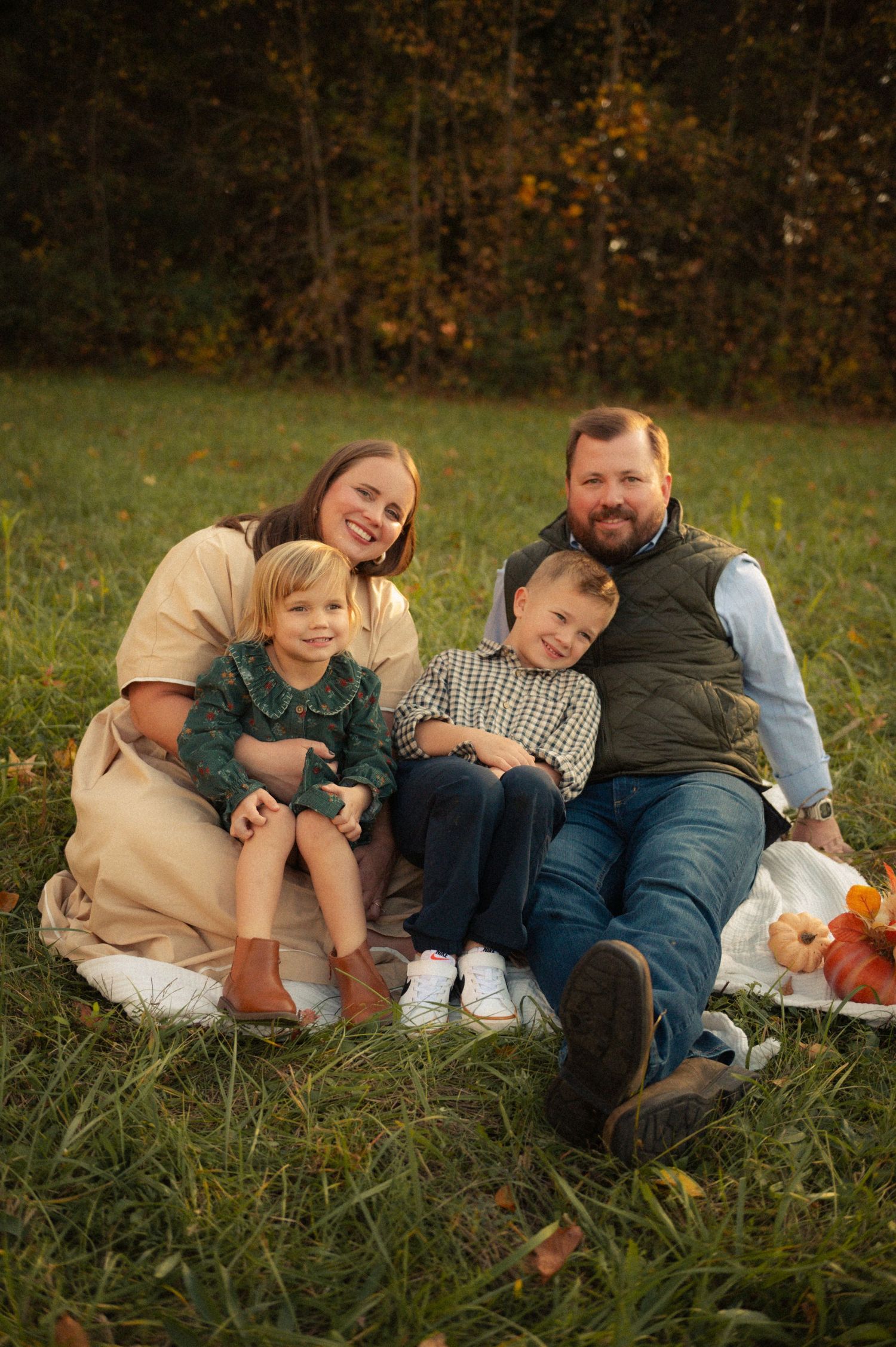 Family of four smiling together on a blanket in a green autumn field, enjoying a cozy outdoor fall photo session.