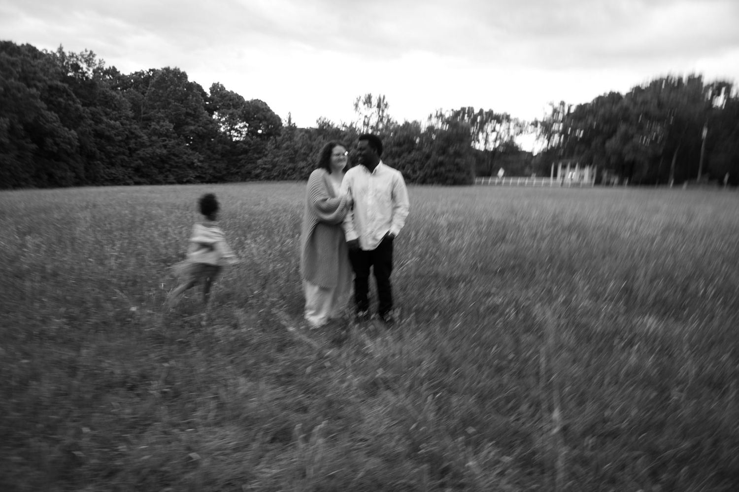 Black and white photo of a couple standing in a grassy field with a child running nearby, trees in background.