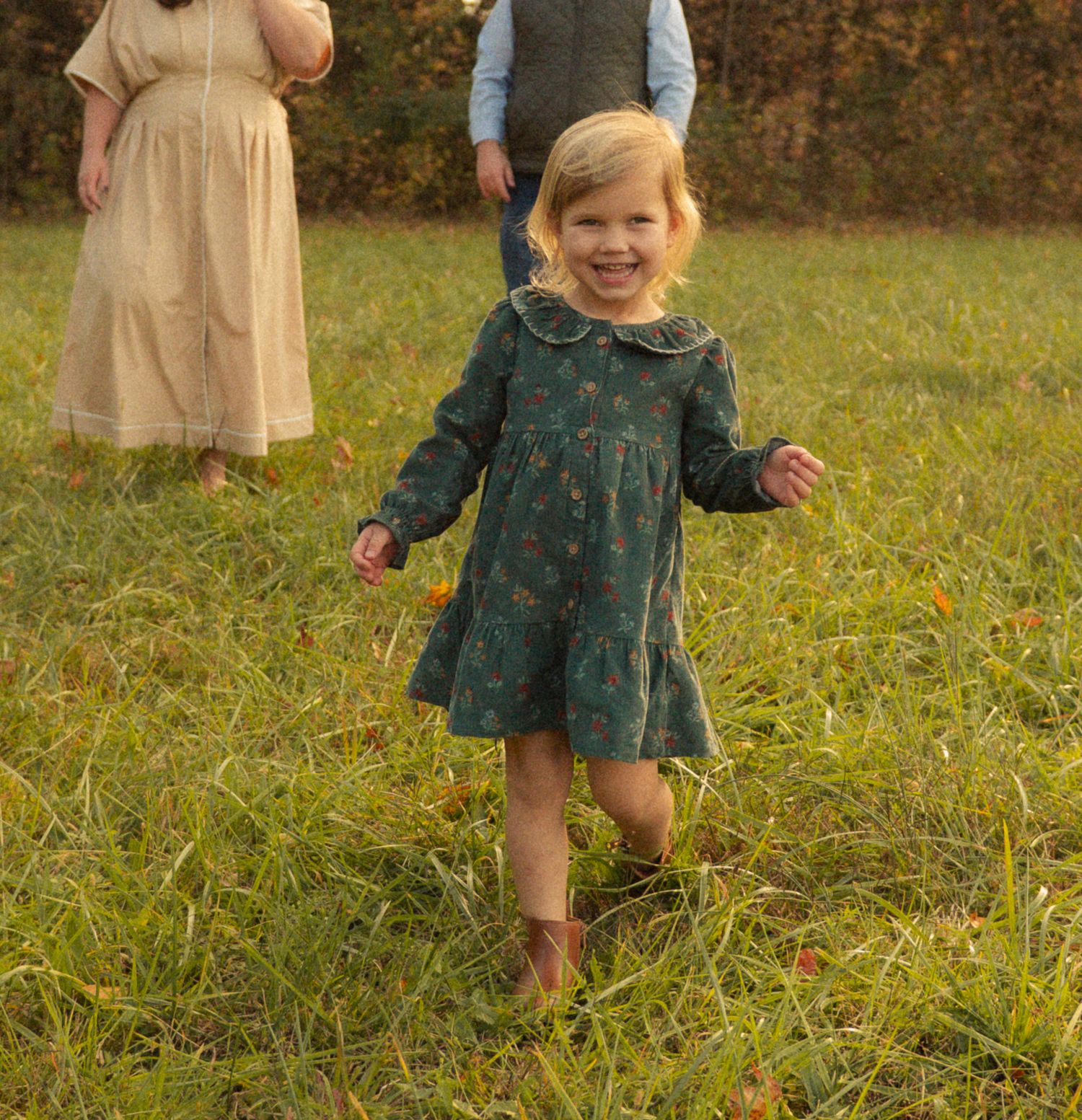 Smiling toddler girl in green floral dress running barefoot through a grassy field with adults standing behind her.