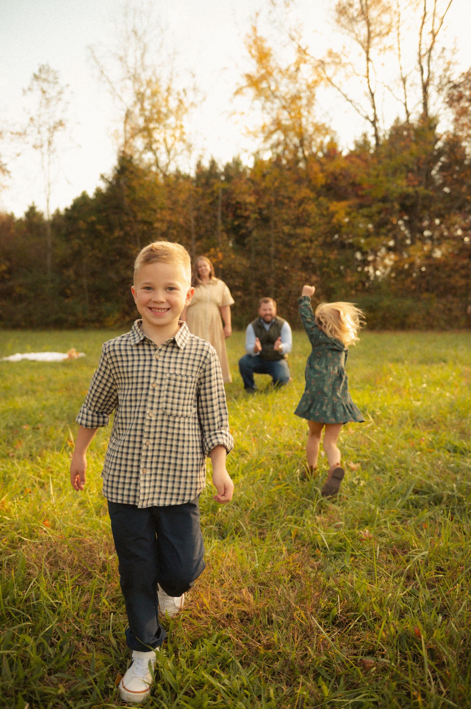 A smiling boy in a plaid shirt walks toward the camera as his family plays in a sunlit green field with autumn trees.