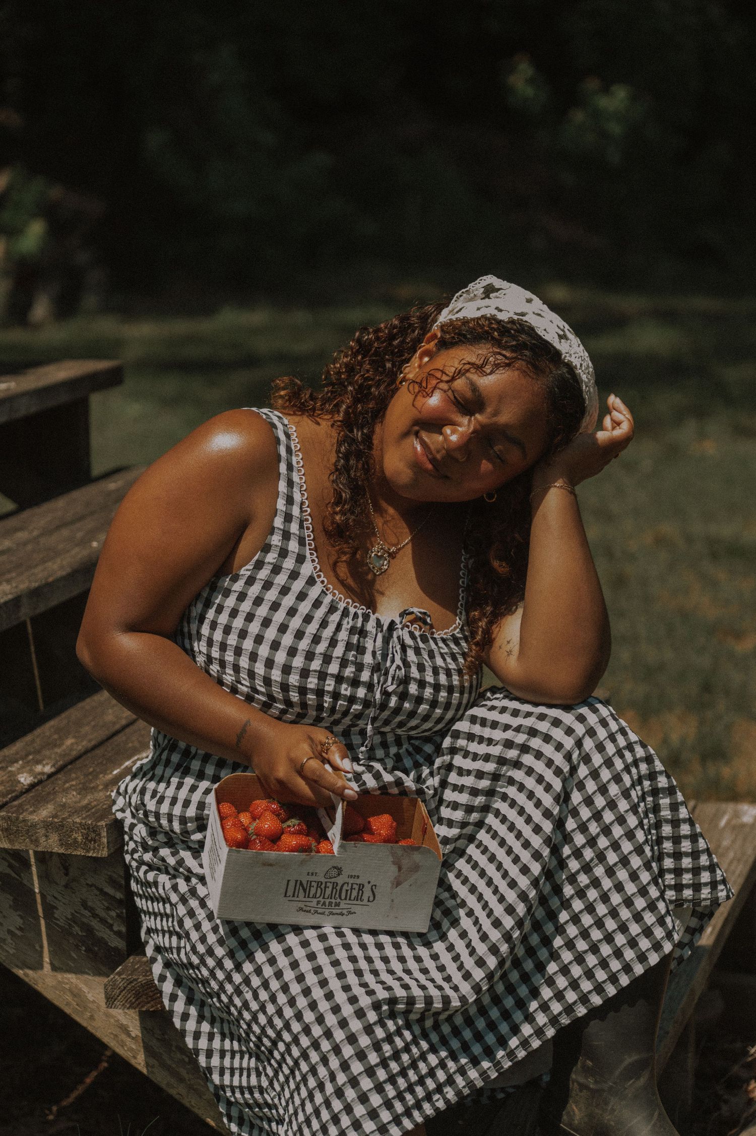 Woman in gingham dress smiling outdoors, holding a box of strawberries from Lindenbrook's Farm.