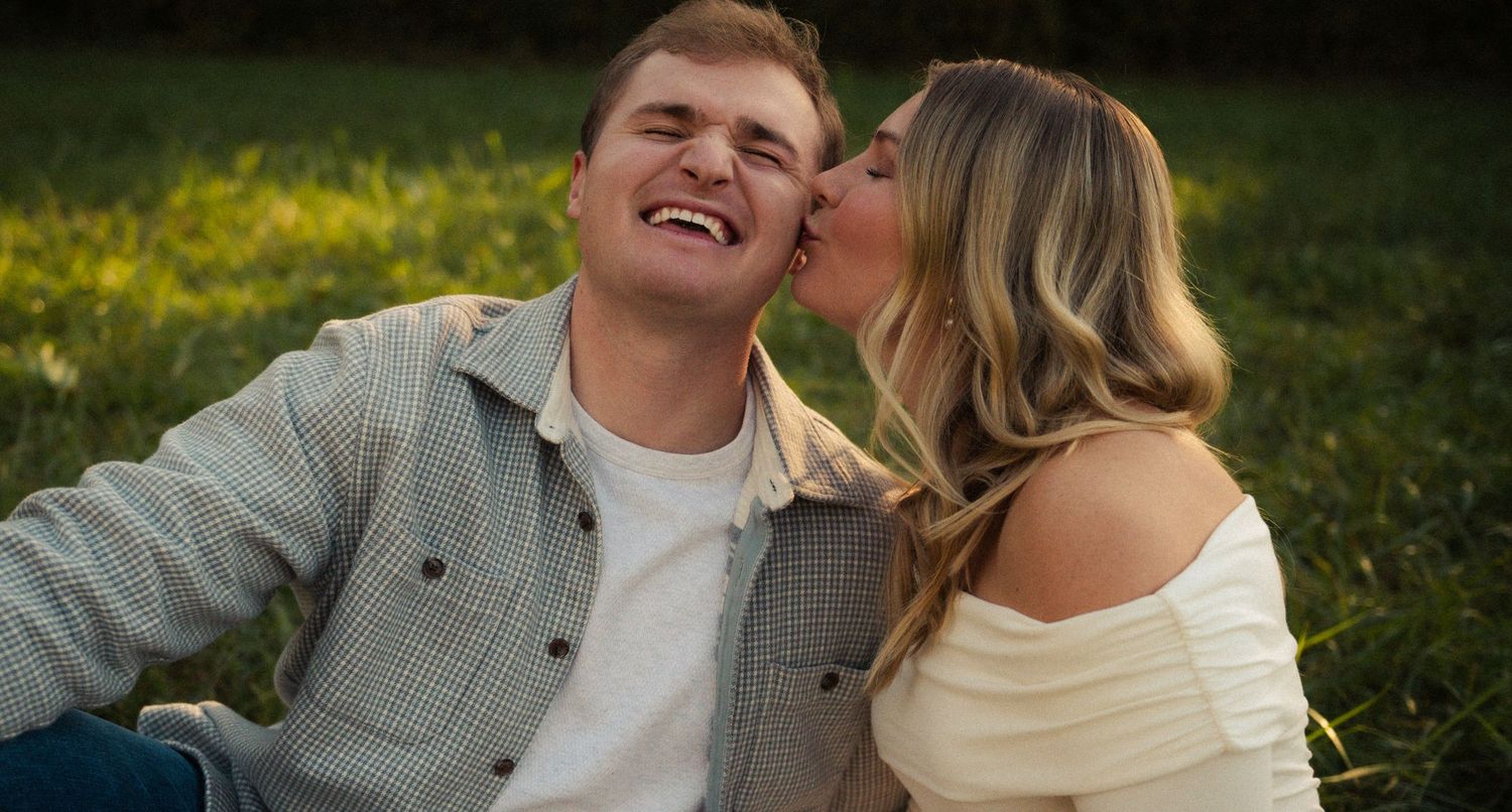 A laughing man receives a kiss on the cheek from a smiling blonde woman while sitting on green grass outdoors.