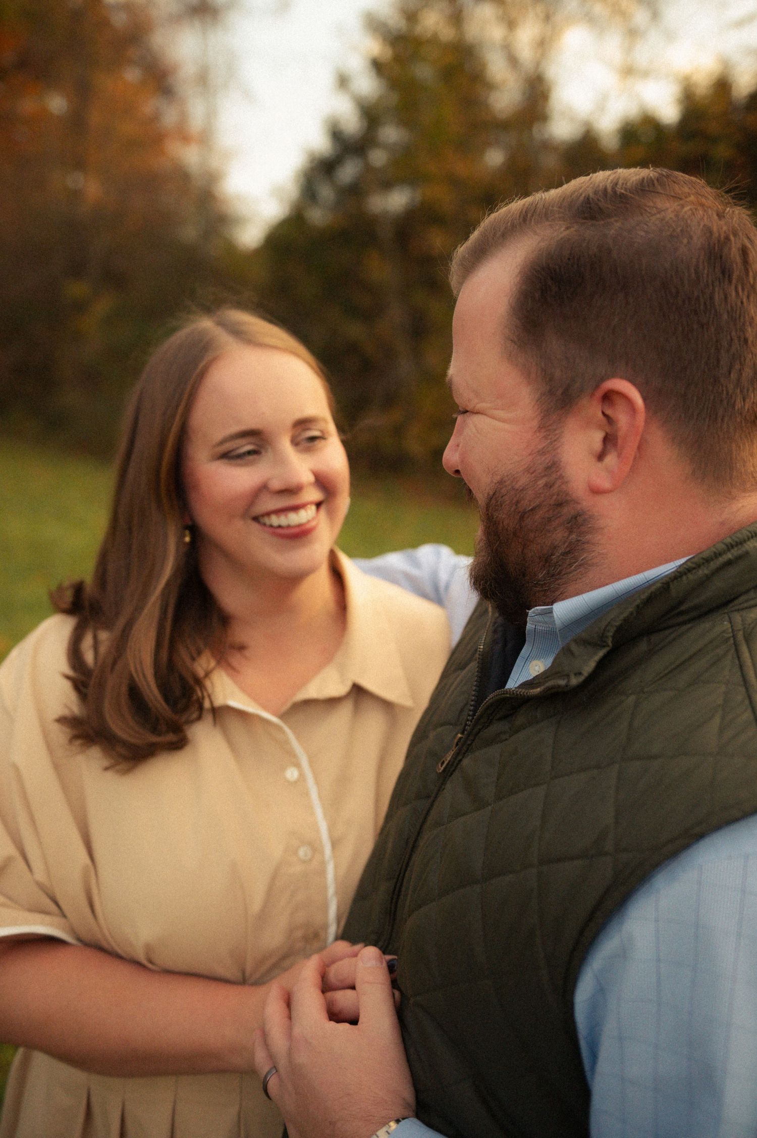 A smiling couple holds hands outdoors in autumn, woman in beige dress, man in green vest and blue shirt.
