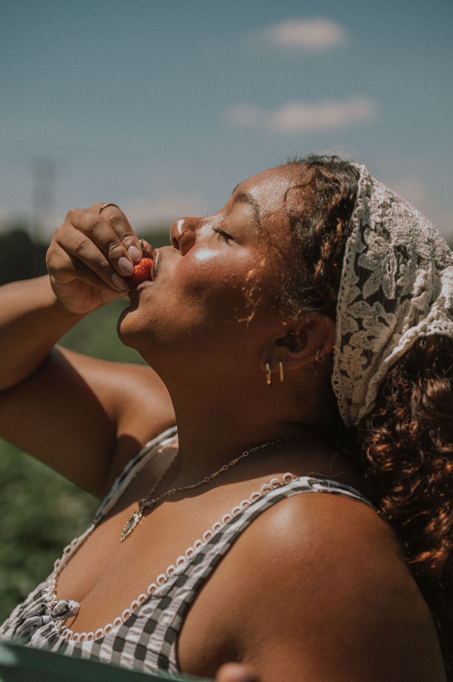 Woman tilting head back eating a cherry outdoors on a sunny day, wearing a floral headscarf and gingham top.