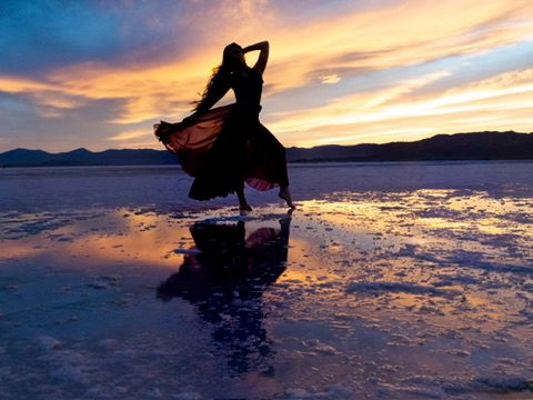Silhouette of a woman in a flowing dress dancing on a reflective salt flat at sunset.