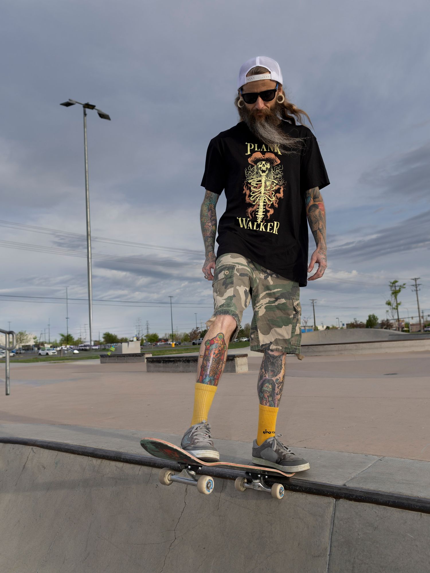 Tattooed skateboarder in camo shorts and black tee performing trick at outdoor skate park.