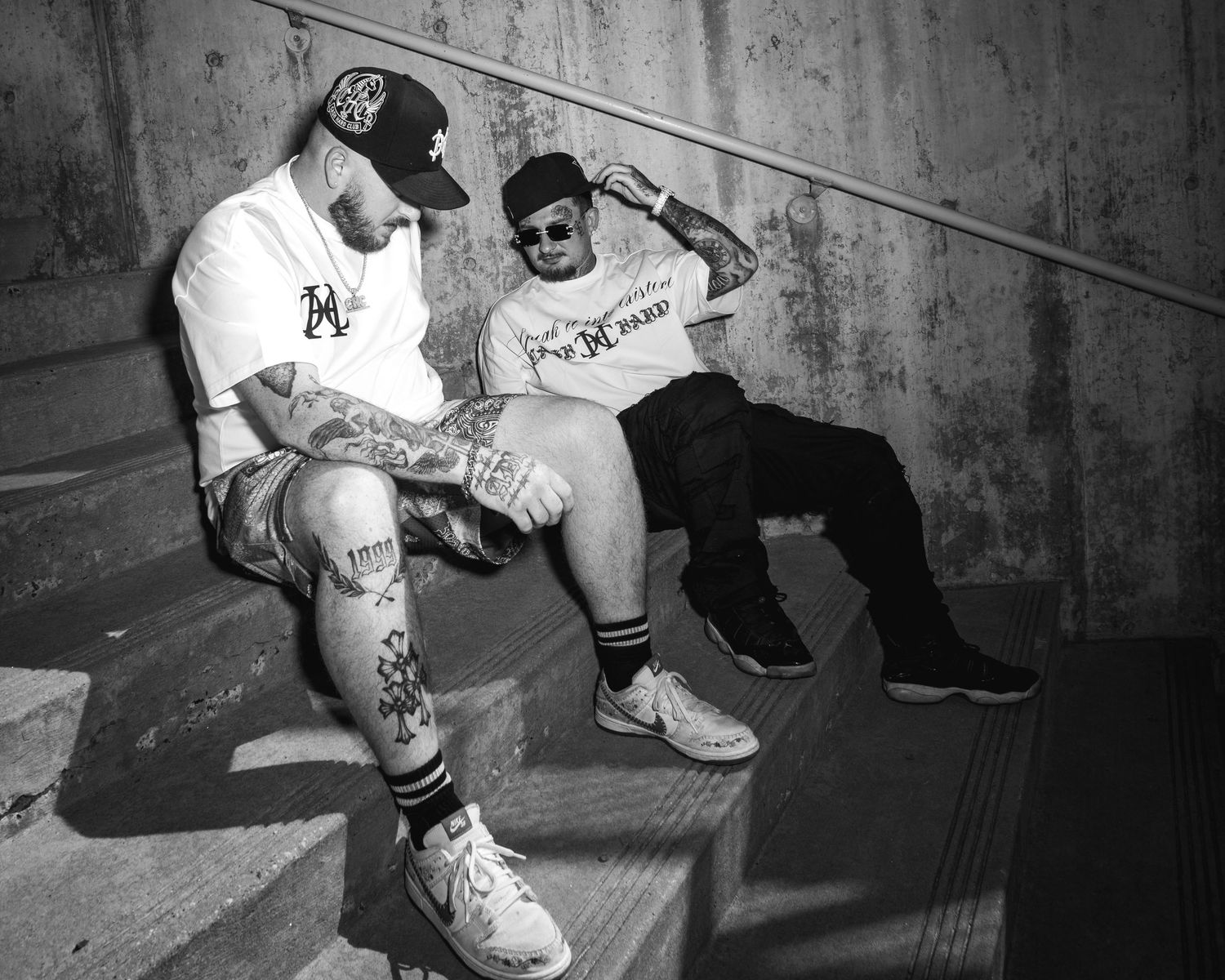 Two tattooed men in streetwear sit on concrete steps in a black and white urban photo.
