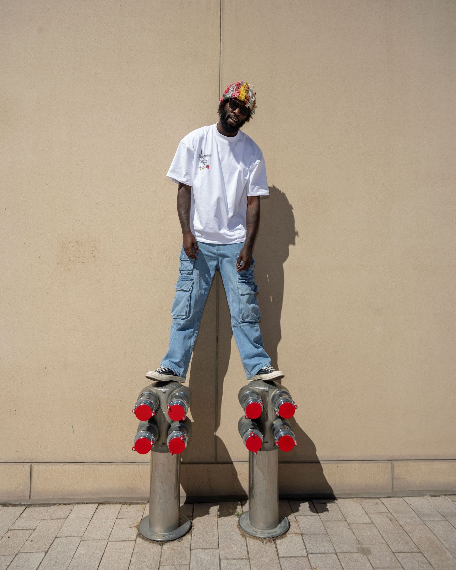 Man in white tee, cargo jeans, and colorful cap standing on two metal bollards with red caps.