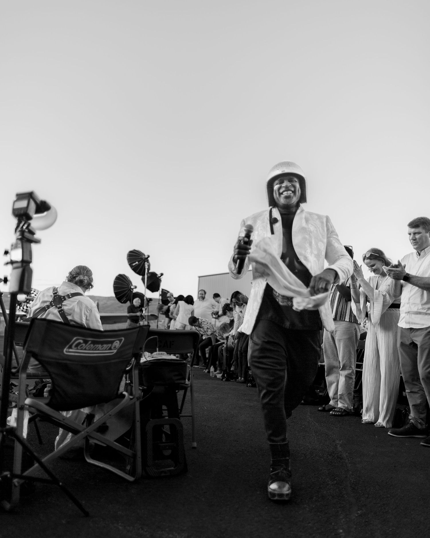 Black and white fashion show backstage with model in white jacket and helmet walking among crew and equipment.