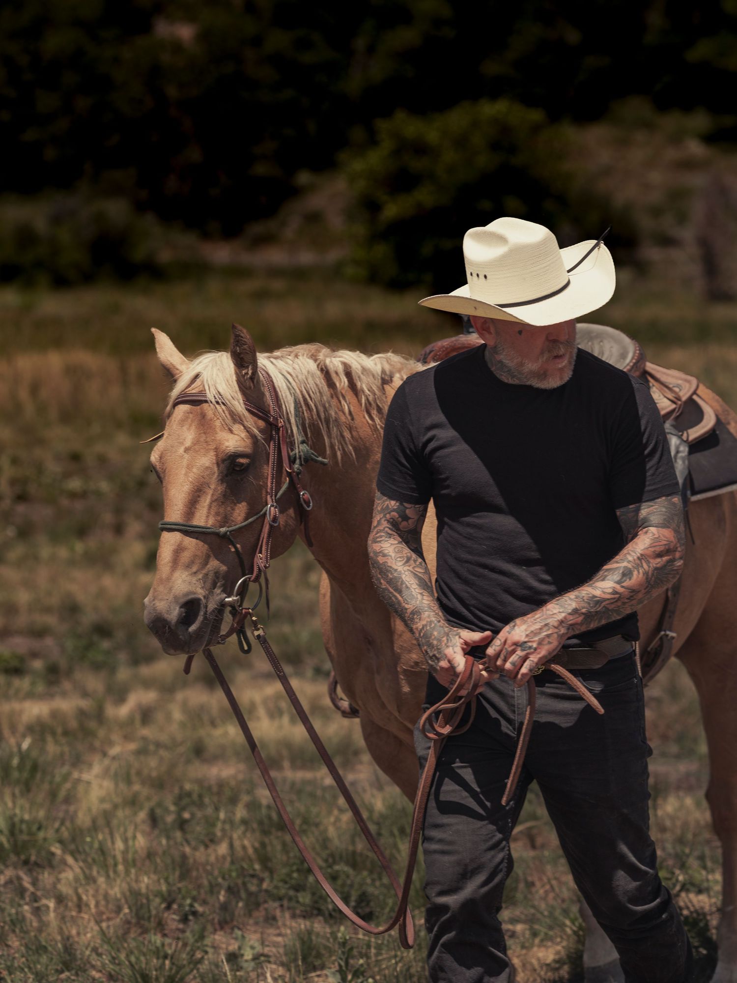 Tattooed man in cowboy hat leading a palomino horse through a grassy field outdoors.