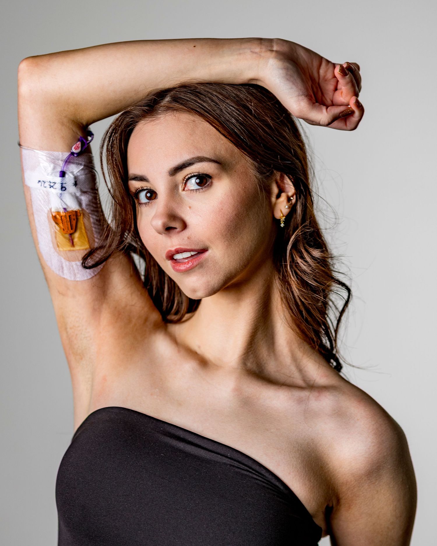 Young woman in black strapless top posing with arm raised, showing a medical IV or patch on her upper arm.