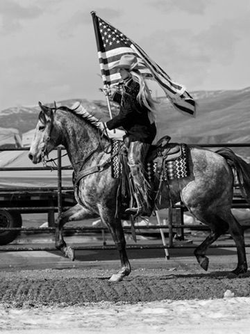 Black and white photo of rider on grey horse carrying American flag at rodeo.