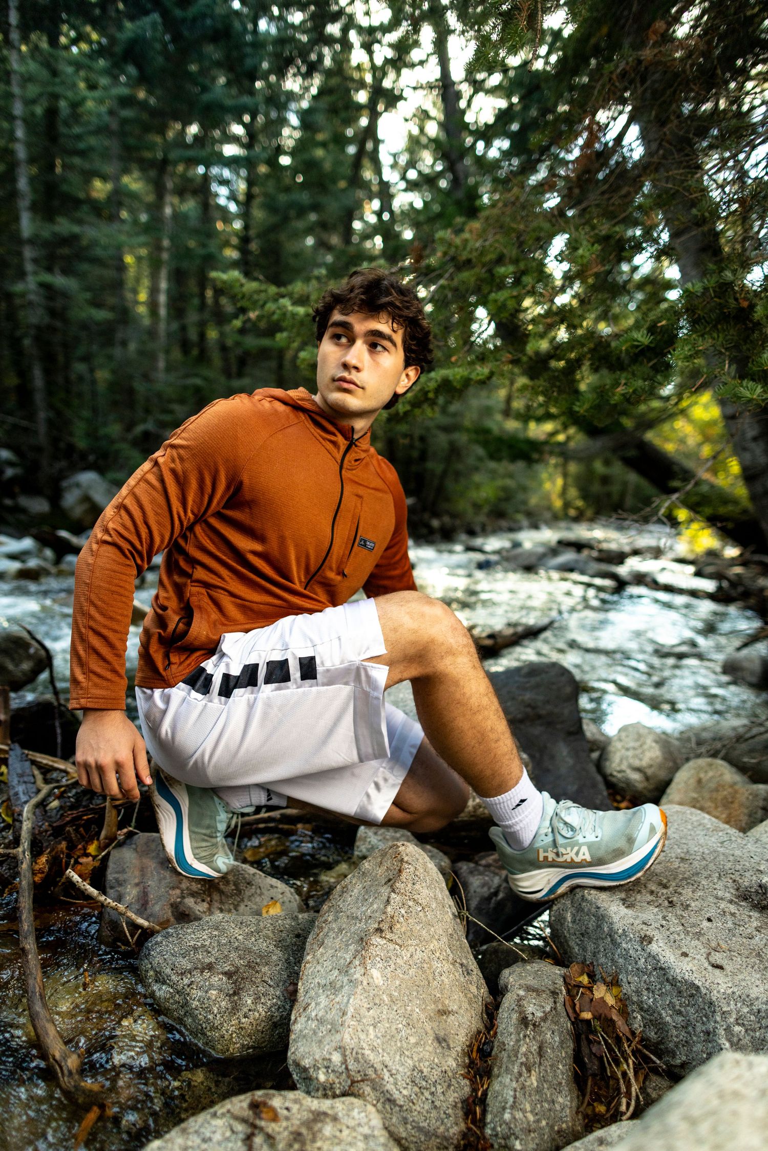 Young man in orange hoodie and white shorts sitting on rocks by a forest stream wearing teal sneakers.