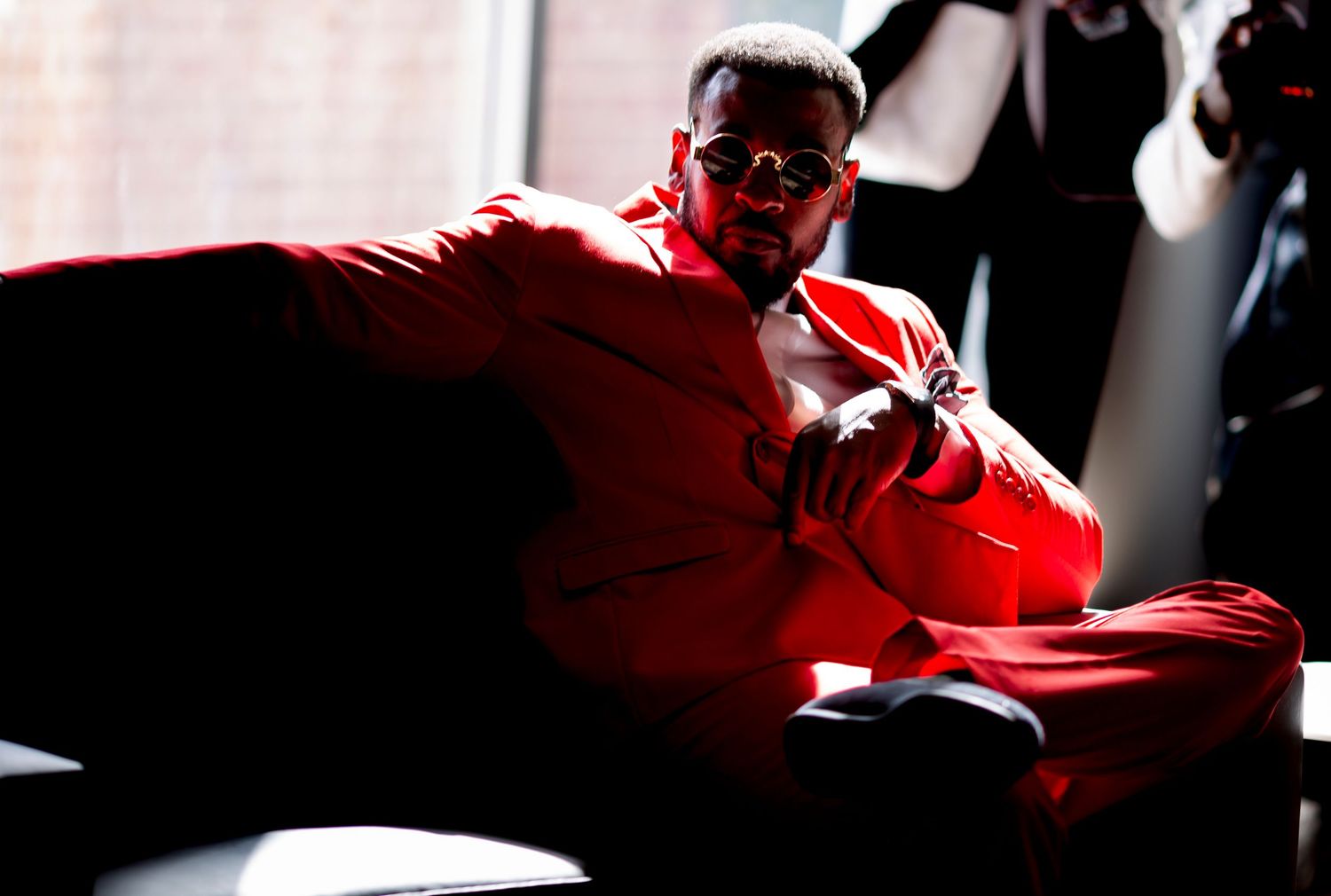 Man in red suit and sunglasses posing confidently on a dark sofa near a window in dramatic lighting.