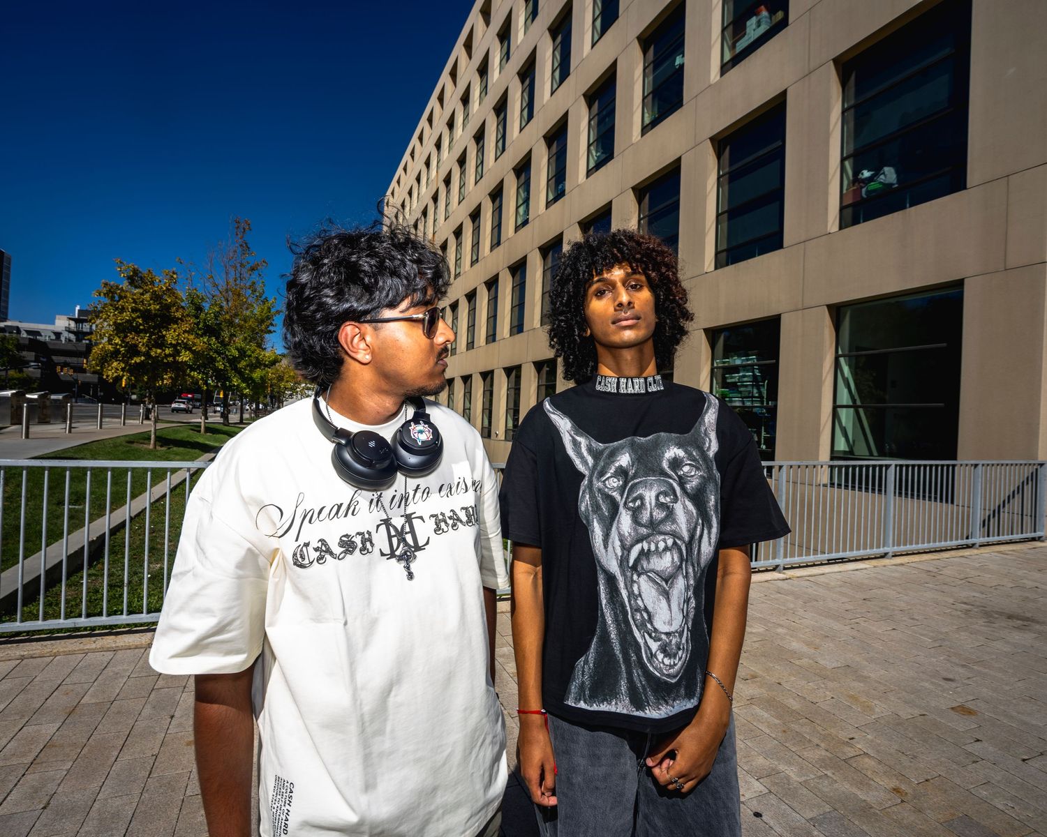 Two young men in graphic tees pose outdoors near a modern building on a sunny day.