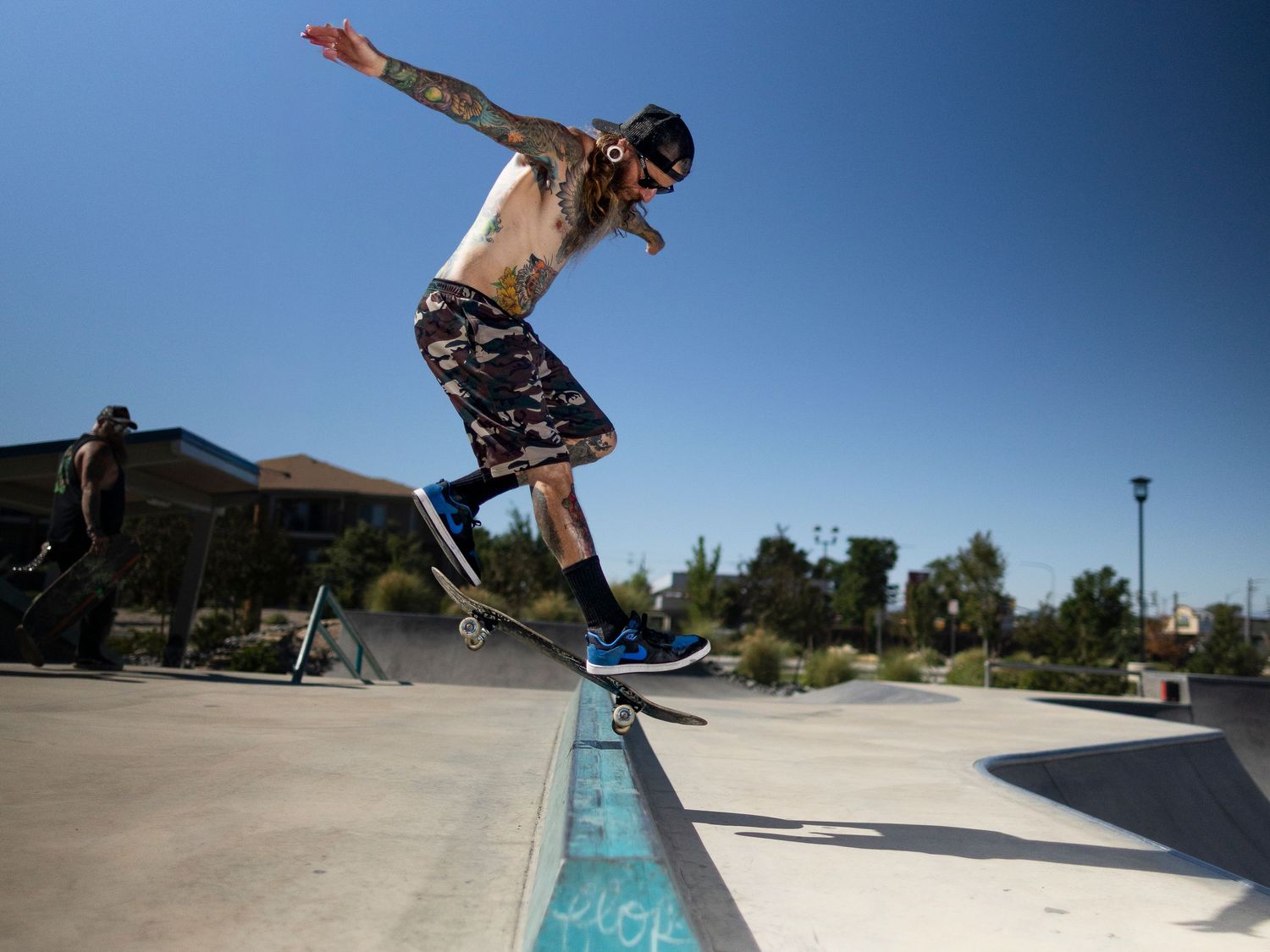Tattooed skateboarder performing a trick on a ramp at an outdoor skate park on a sunny day.