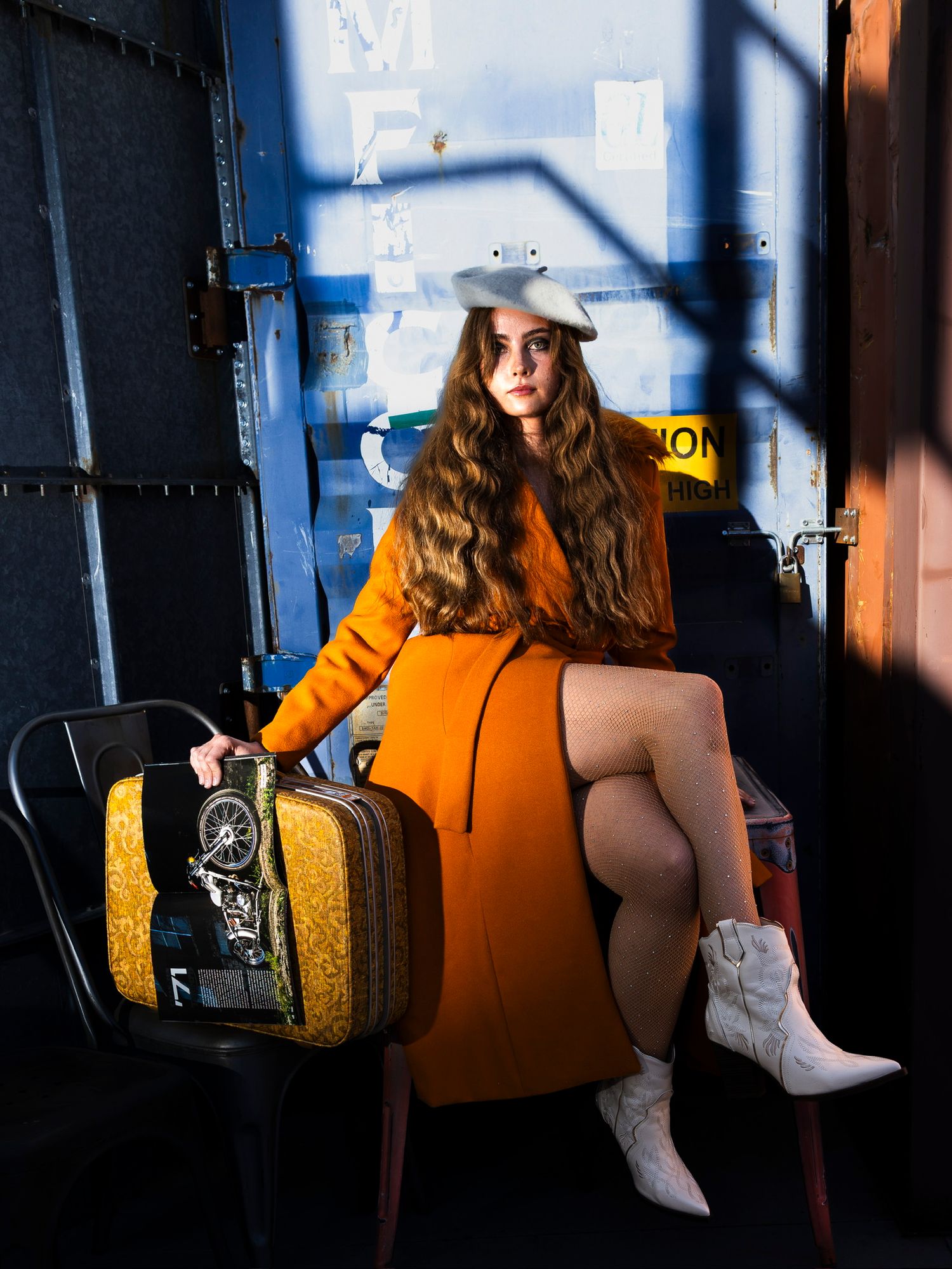 Young woman in orange dress and white beret sits with vintage suitcase in urban setting.