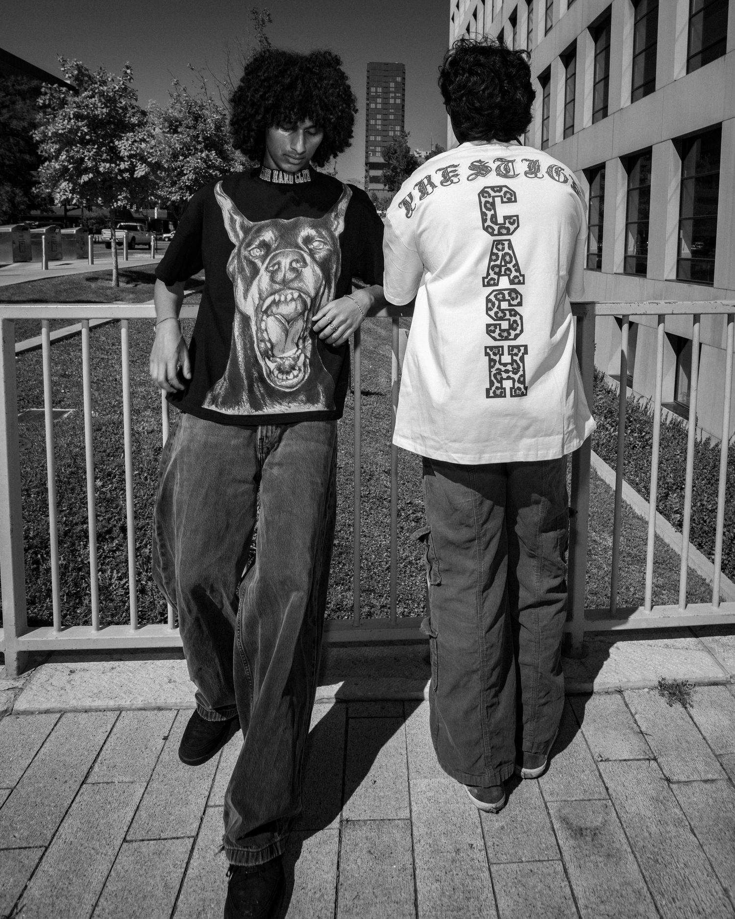 Two young men in streetwear pose outdoors, one wearing a dog graphic tee, the other a white Cash logo shirt.