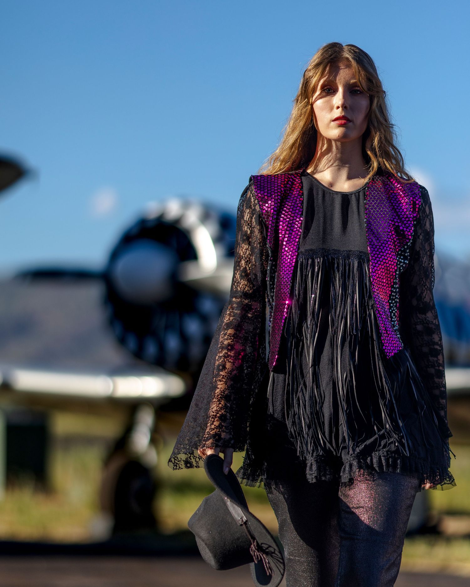 Woman in purple sequin fringe jacket poses before vintage aircraft propeller on sunny airfield.