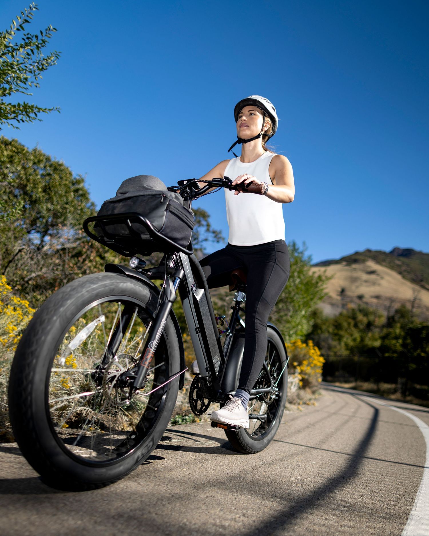 Woman in white tank top and helmet stands with black fat-tire electric bike on sunny road with hills.