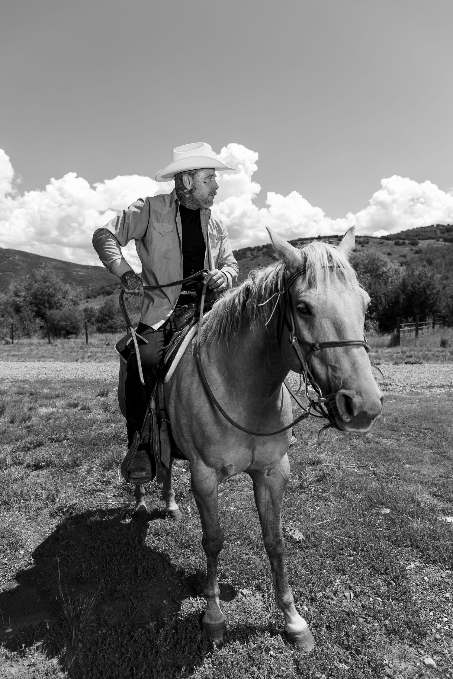Black and white portrait of cowboy on horseback against cloudy sky in rural landscape.