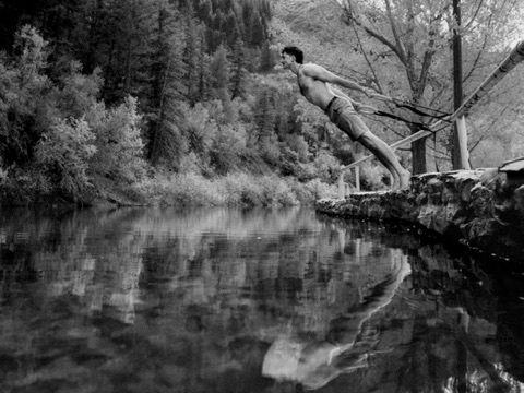 Man diving into calm reflective mountain river surrounded by forest in black and white photo.