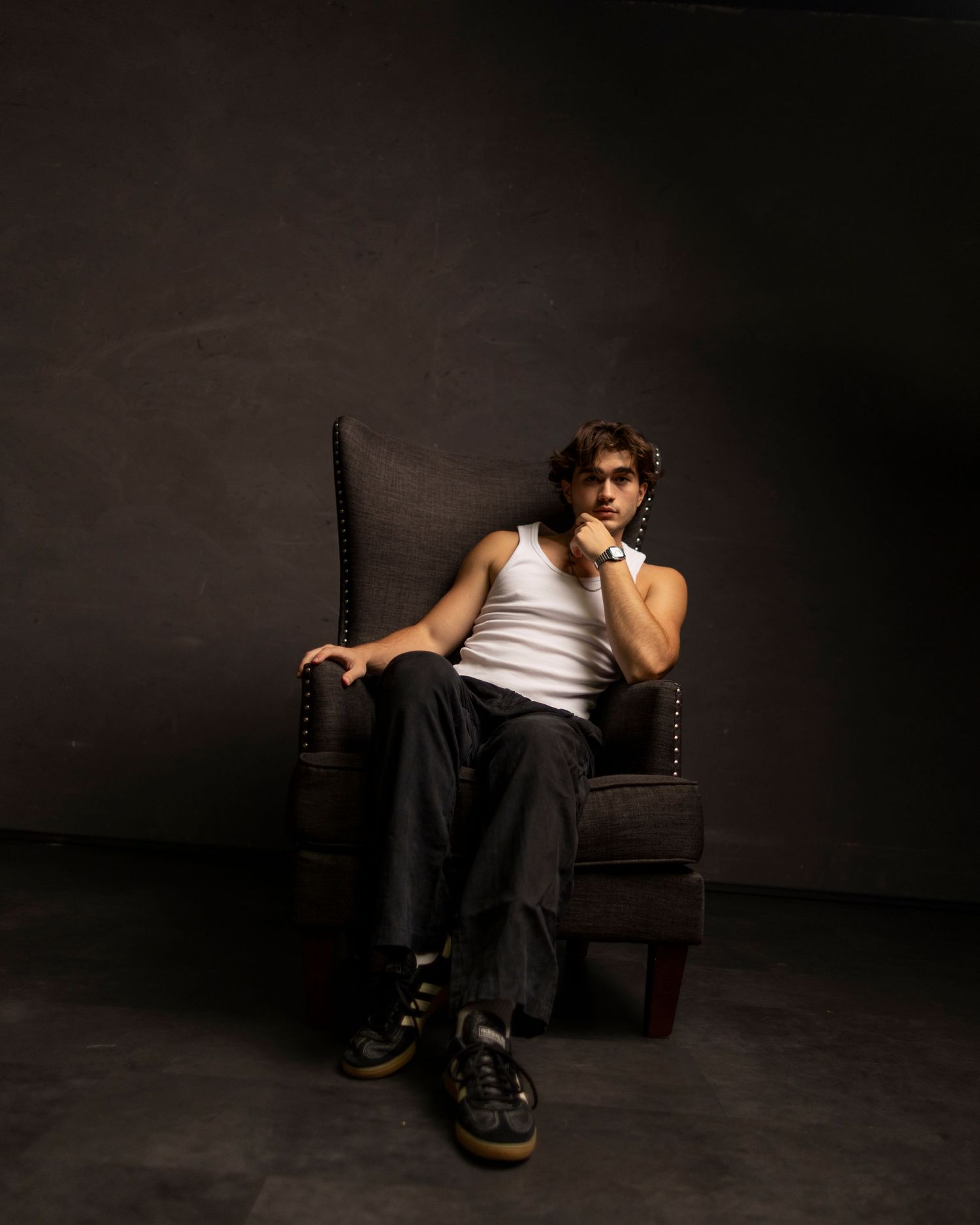 Young man in white tank top and dark pants sits confidently in an armchair against a dark studio background.