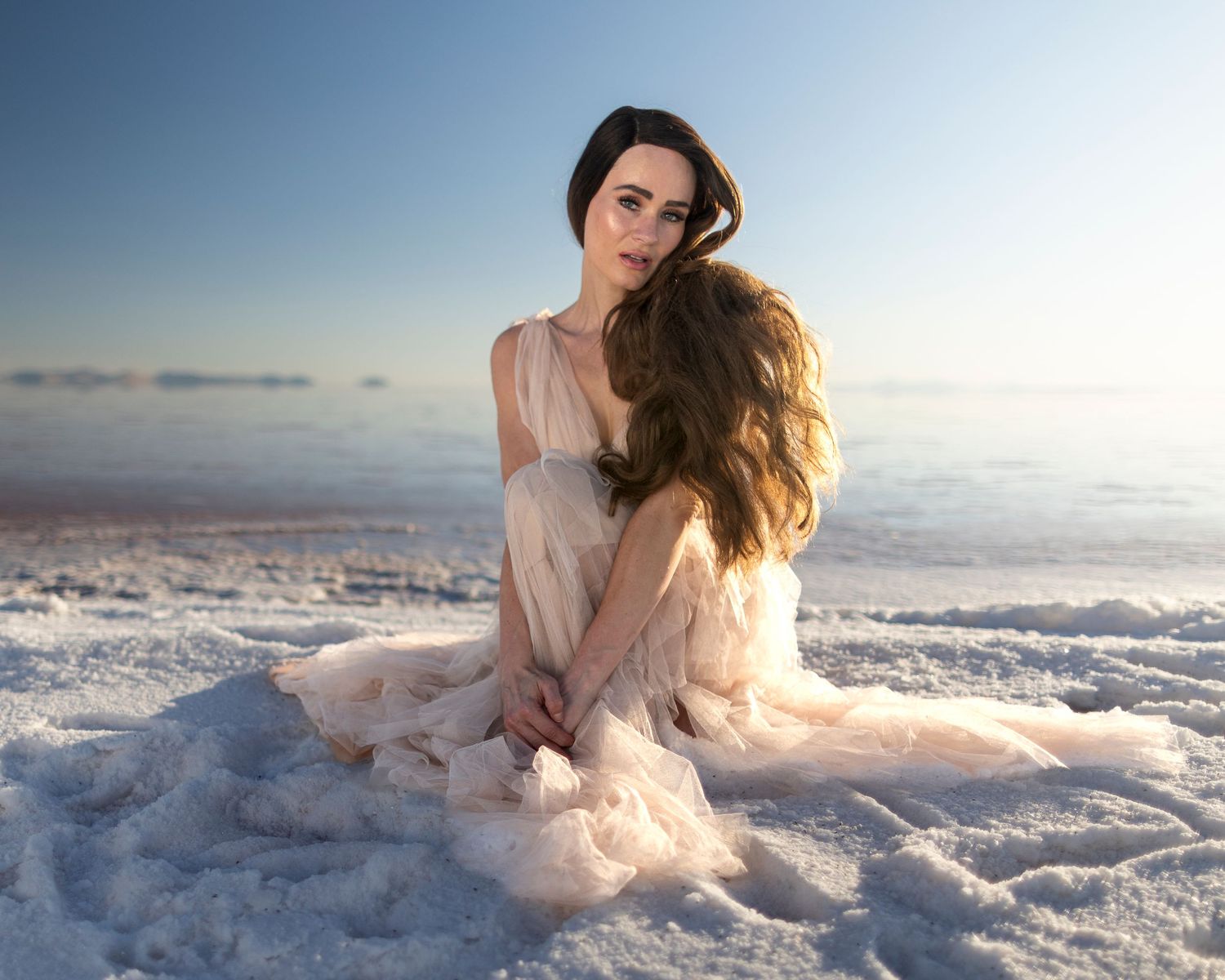 A young woman in a flowing blush gown sits gracefully on white salt flats under a clear blue sky.