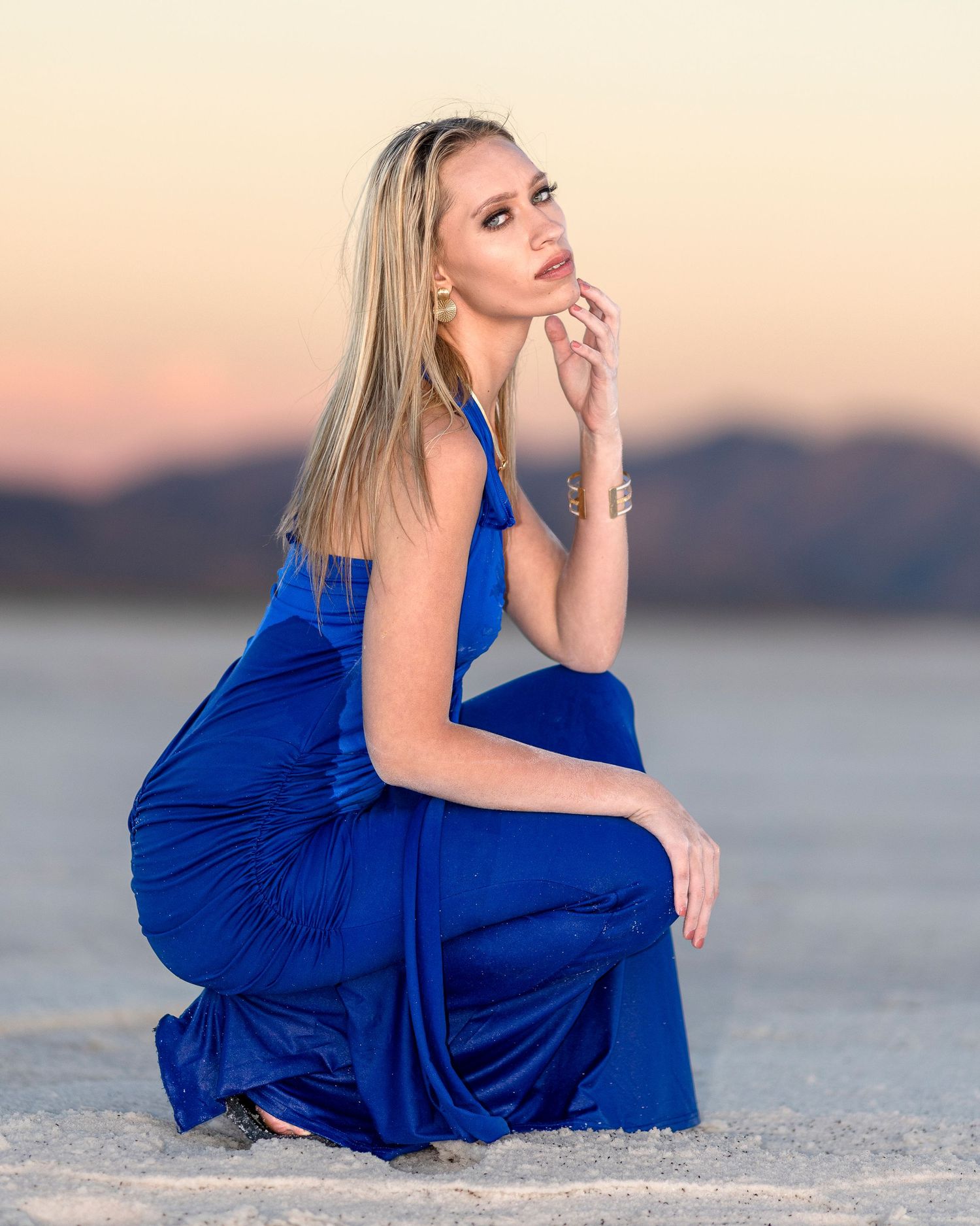 Blonde woman in a royal blue pleated gown crouching gracefully on white salt flats at sunset.